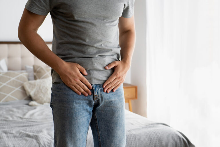 Person stands in a bedroom, hands pressed to the lower abdomen, suggesting stomach pain or discomfort in casual clothing (gray tee, jeans).