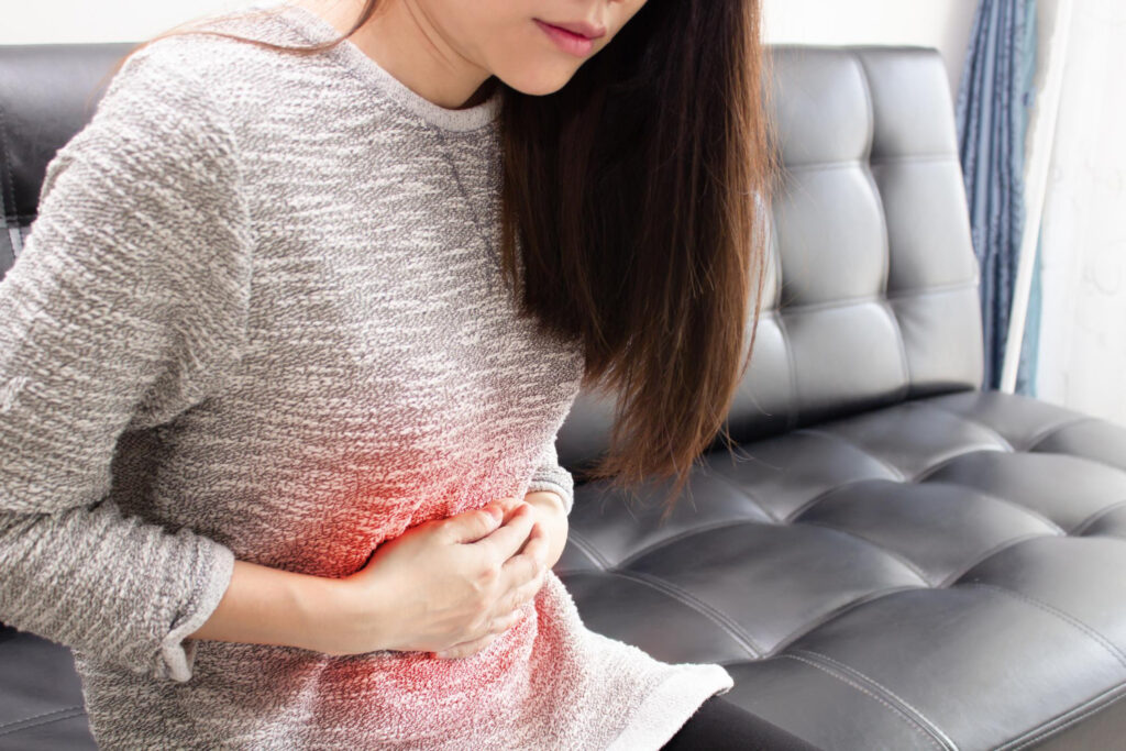 Woman sitting on a gray leather couch, clutching her abdomen in pain.