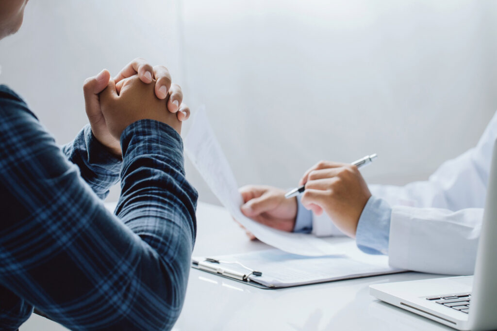 Person in a plaid shirt and a clinician sit at a desk; hands clasped in a gesture of agreement during a medical consultation.